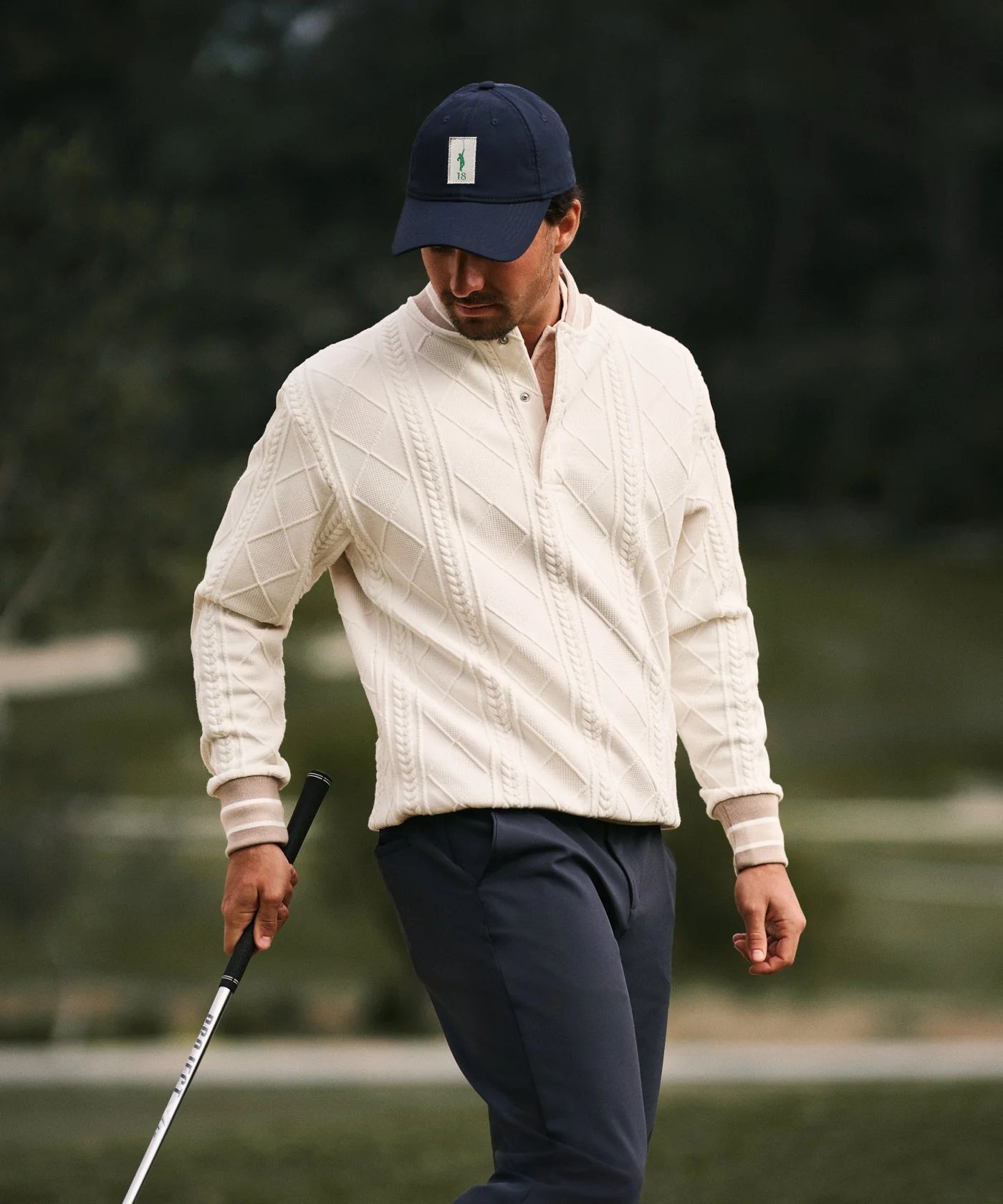 Golfer in a natural cream cable-knit quarter-zip, navy cap and navy trousers.