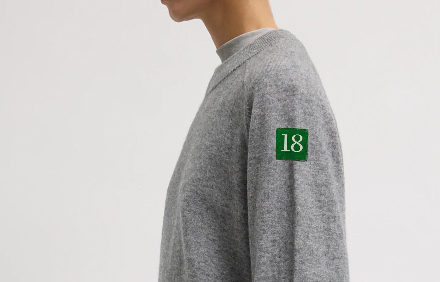 Heather gray cashmere crewneck with a felted green 18 patch on the upper arm.