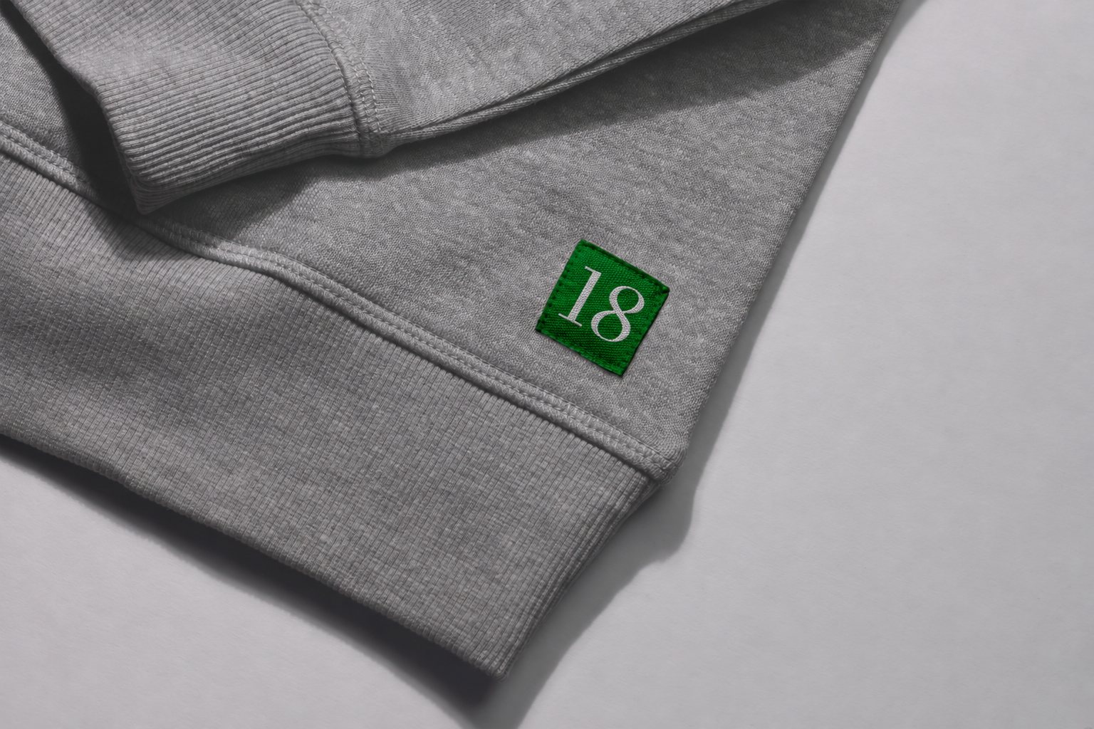 A square green felt 18 patch stitched to the hem of heather gray knitwear.